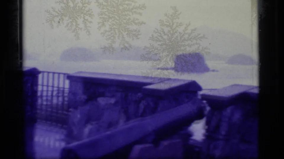 Blue-tinted 1977 Alaska home-movie still with a cannon beside a stone wall overlooking misty coastal water and islands.
