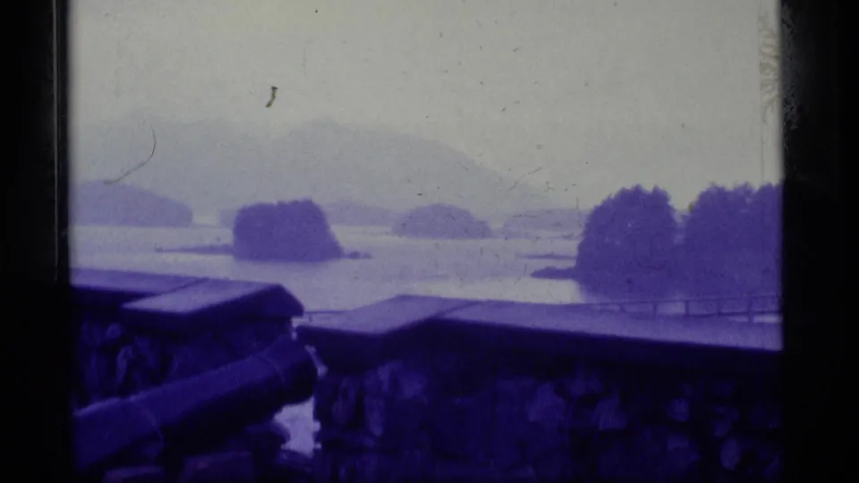 Blue-purple 1977 Alaska 8mm frame of a quiet coastal vista beyond a stone wall, with low islands, haze, and a partial cannon.