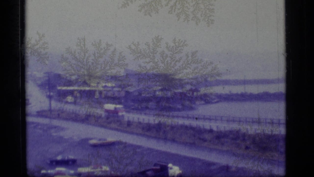 1977 Alaskan town view from above with older cars, low buildings, bare branches, and a hazy waterfront in the distance.
