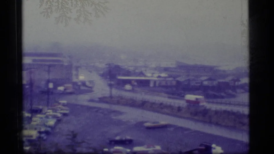 Hazy 1977 Alaska street view with older cars, a red-and-white van, modest buildings, and a distant waterfront.