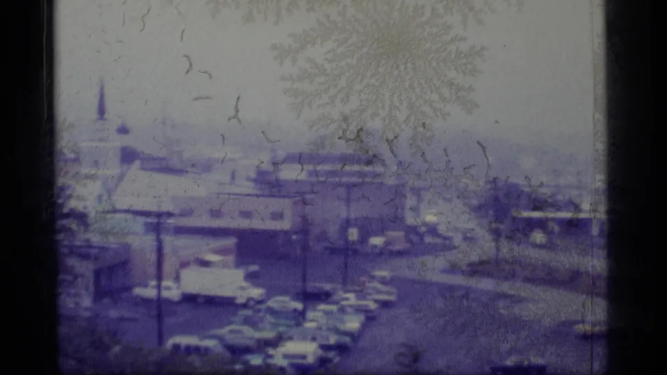 1977 Alaskan town frame with parked cars, low rooftops, bare tree branches, and a church steeple rising through haze.