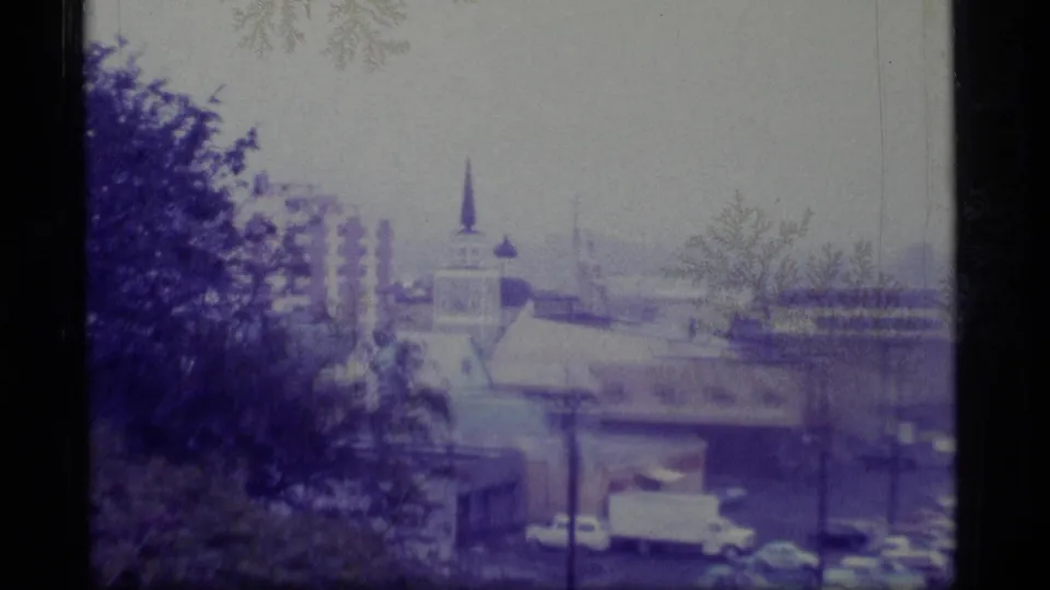 1977 Alaska town view with a church steeple, low buildings, parked cars, trees, and a muted waterfront background.