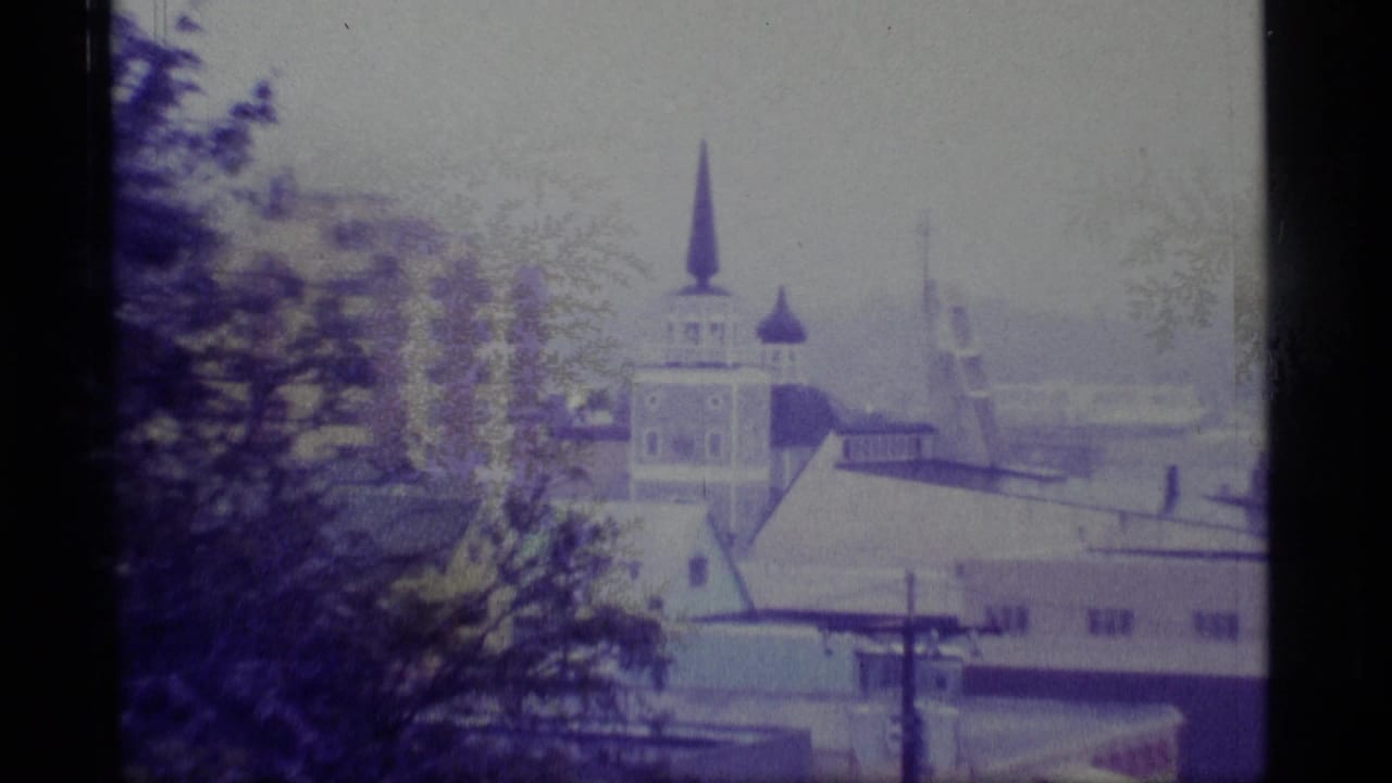 1977 Alaska 8mm frame showing a distant church steeple, low rooftops, trees, and utility poles in an overcast coastal town.