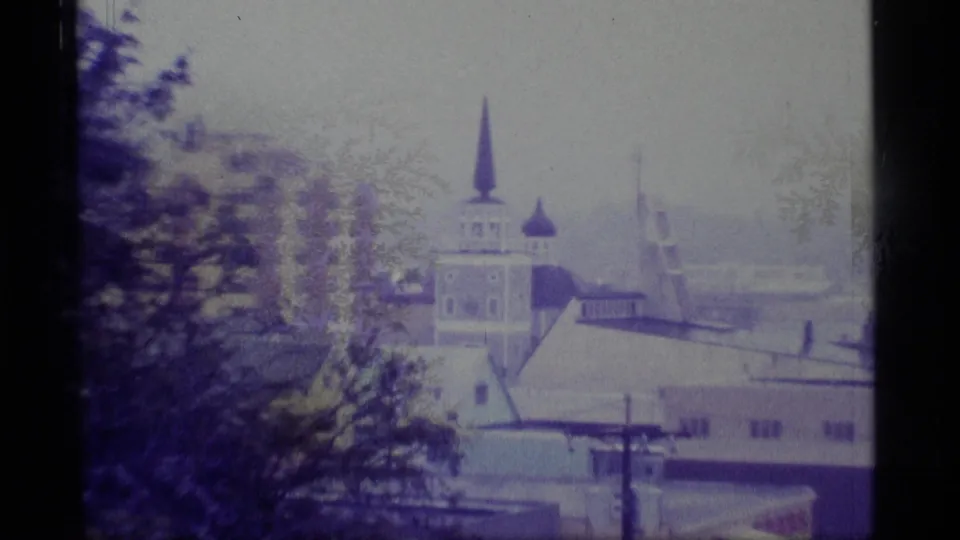 1977 Alaska 8mm frame showing a distant church steeple, low rooftops, trees, and utility poles in an overcast coastal town.