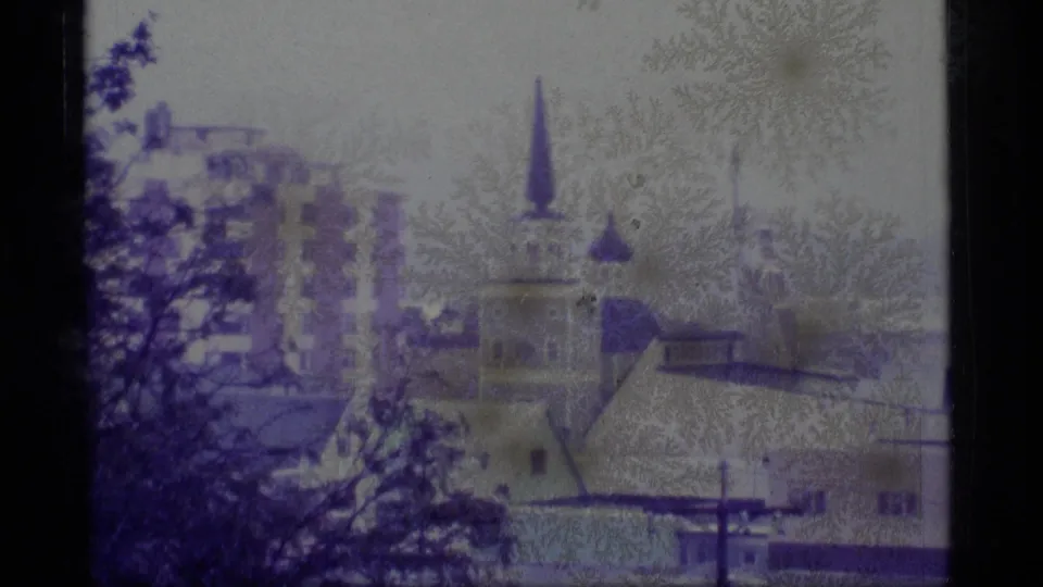 1977 Alaska home-movie frame with a church steeple, clustered buildings, trees, and pale sky, softened by grain and color shift.