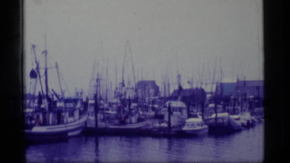1977 Alaska 8mm frame showing a harbor lined with fishing boats, tall masts, docks, waterfront buildings, and reflective water.