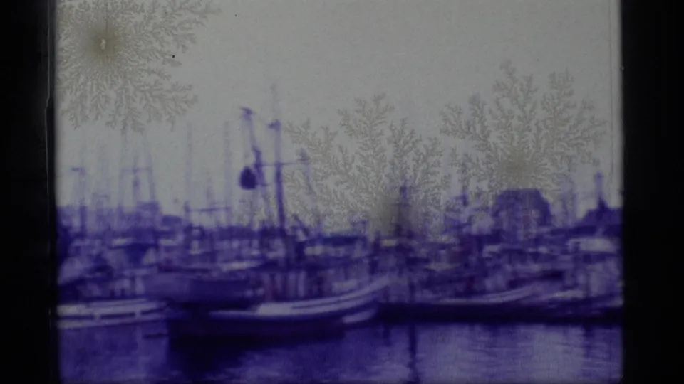 1977 Alaska home-movie frame of a harbor with moored boats, masts, waterfront structures, water, and strong purple film cast.