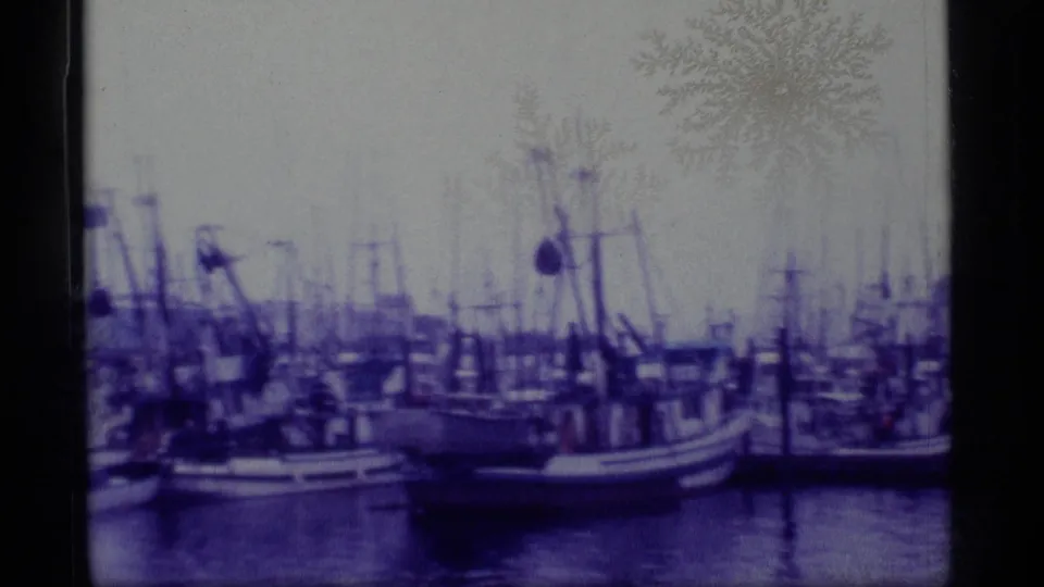 1977 Alaska 8mm frame showing docked fishing boats, masts, rigging, pilings, calm water, and a hazy shoreline.
