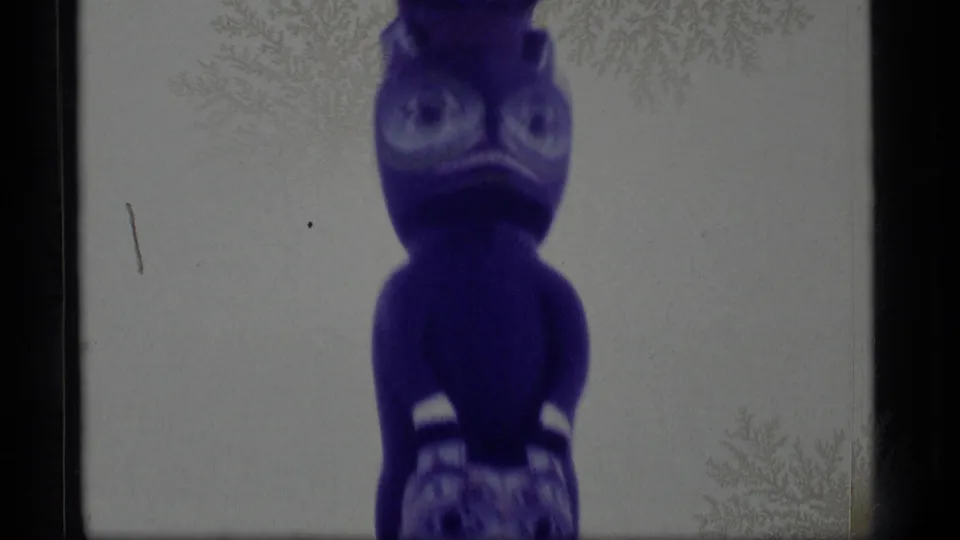 1977 Alaska frame of a purple-tinted carved pole figure with prominent eyes, rounded body form, and visible 8mm film wear.