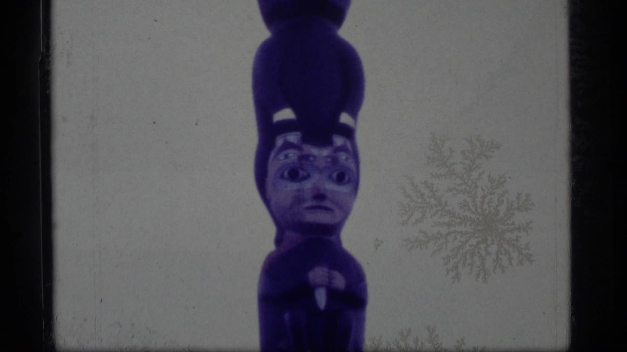 1977 Alaska 8mm frame showing the upper carved faces of a purple-painted totem pole against a pale sky.
