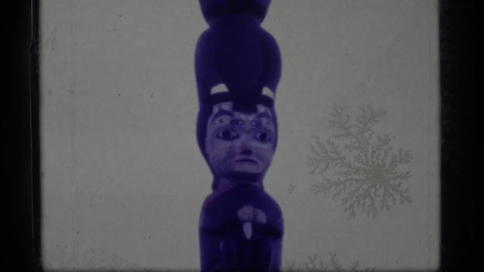 1977 Alaska 8mm frame showing the upper carved faces of a purple-painted totem pole against a pale sky.