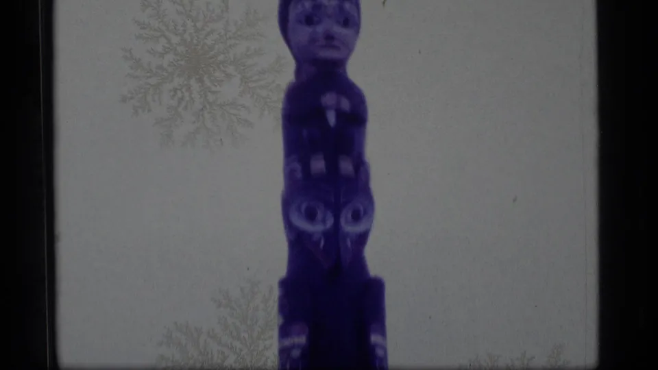 1977 Alaska 8mm frame with the painted totem pole centered, showing a face above bird-like carved forms.