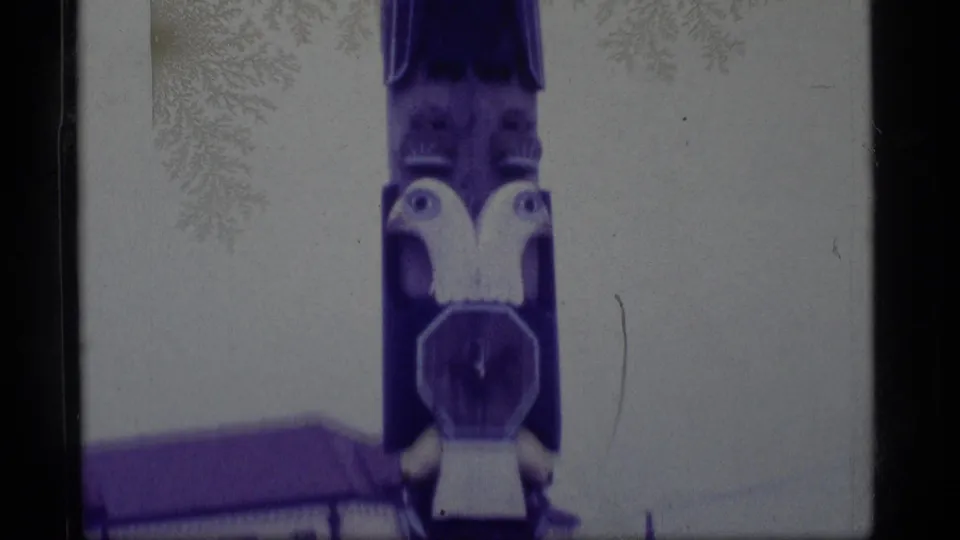 1977 Alaska 8mm frame showing the lower painted pole, an owl-like carving, and a low building roof in the background.