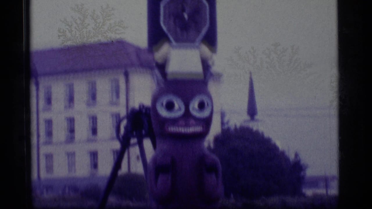 1977 Alaska Super 8 frame showing a wide-eyed totem figure before a pale building, with soft focus and purple color shift.