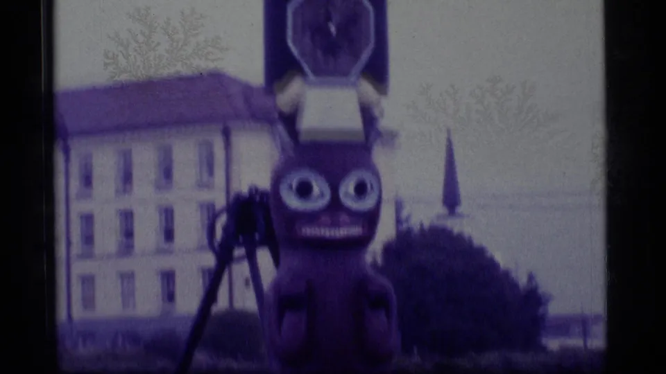 1977 Alaska Super 8 frame showing a wide-eyed totem figure before a pale building, with soft focus and purple color shift.