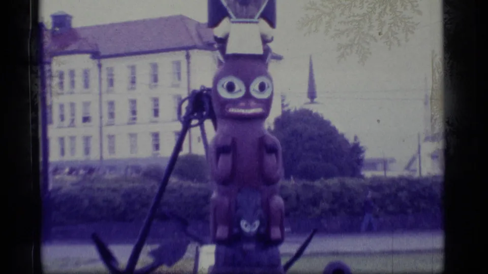 1977 Alaska frame of the totem display with a dark anchor nearby, a white building behind, and a distant spire visible.