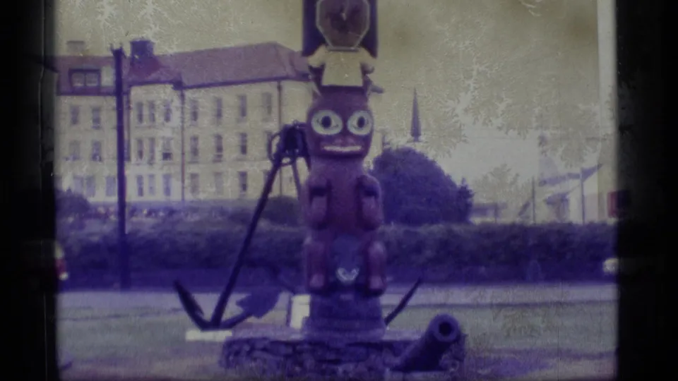 1977 Alaska Super 8 view of the totem display, anchor, cannon, hedge, and pale buildings in a public outdoor setting.