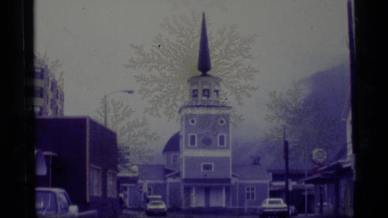 1977 Alaska 8mm frame showing a quiet town street with a tall church steeple, parked cars, storefronts, and bare trees under gray light.