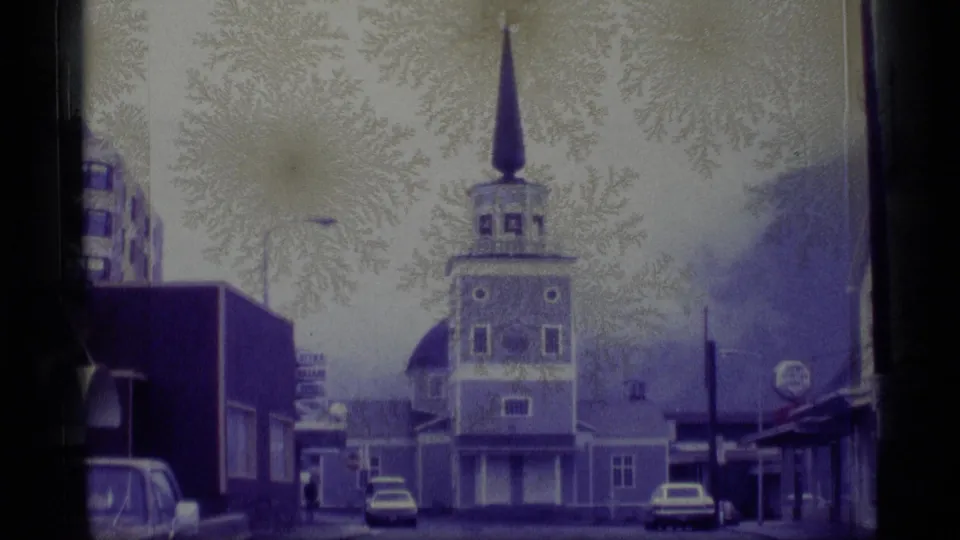 1977 Alaska home-movie frame centered on a church tower along a quiet street with parked cars, nearby buildings, and bare trees.