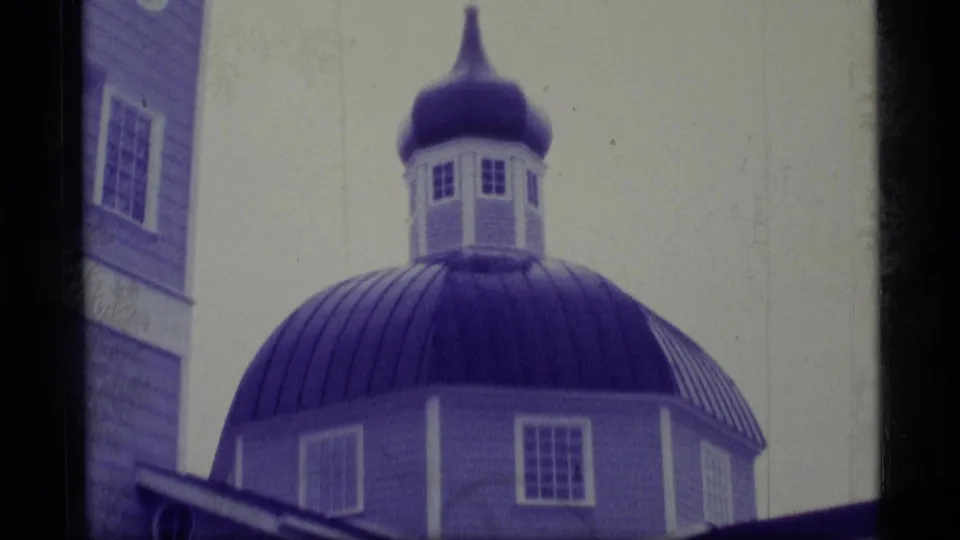 1977 Alaska 8mm frame showing the blue domed church exterior, cupola windows, wall edge, and snowy pale sky.