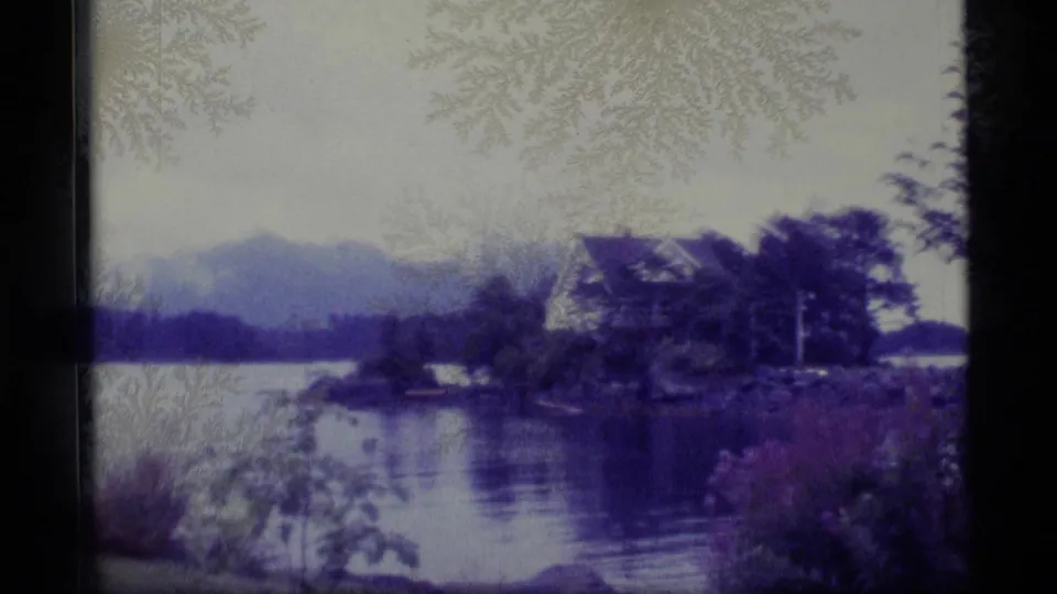 1977 Alaska 8mm frame showing a lakeside house or building, shoreline trees, calm water, and distant mountains.