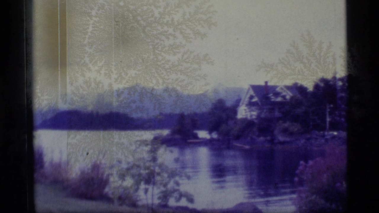 1977 Alaska 8mm frame showing a calm lake, shoreline trees, a waterfront house, and mountains in soft focus.