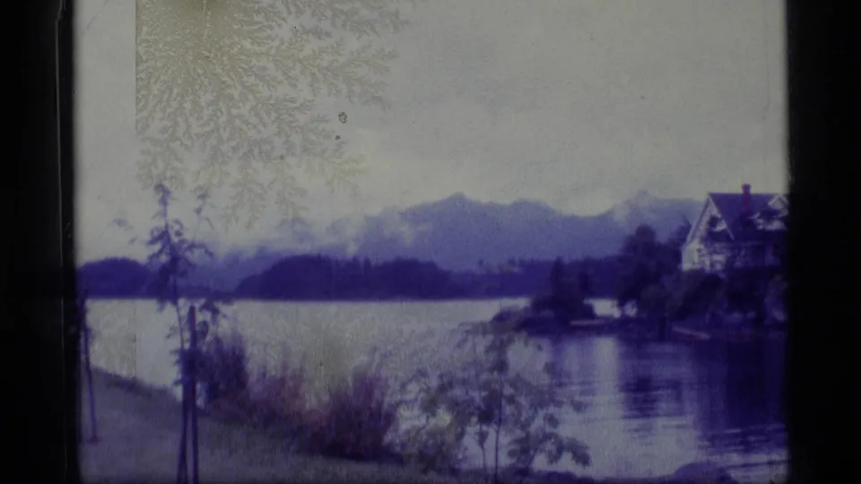 1977 Alaska travel footage frame with a lake, evergreen shoreline, distant mountains, and a partially visible house.