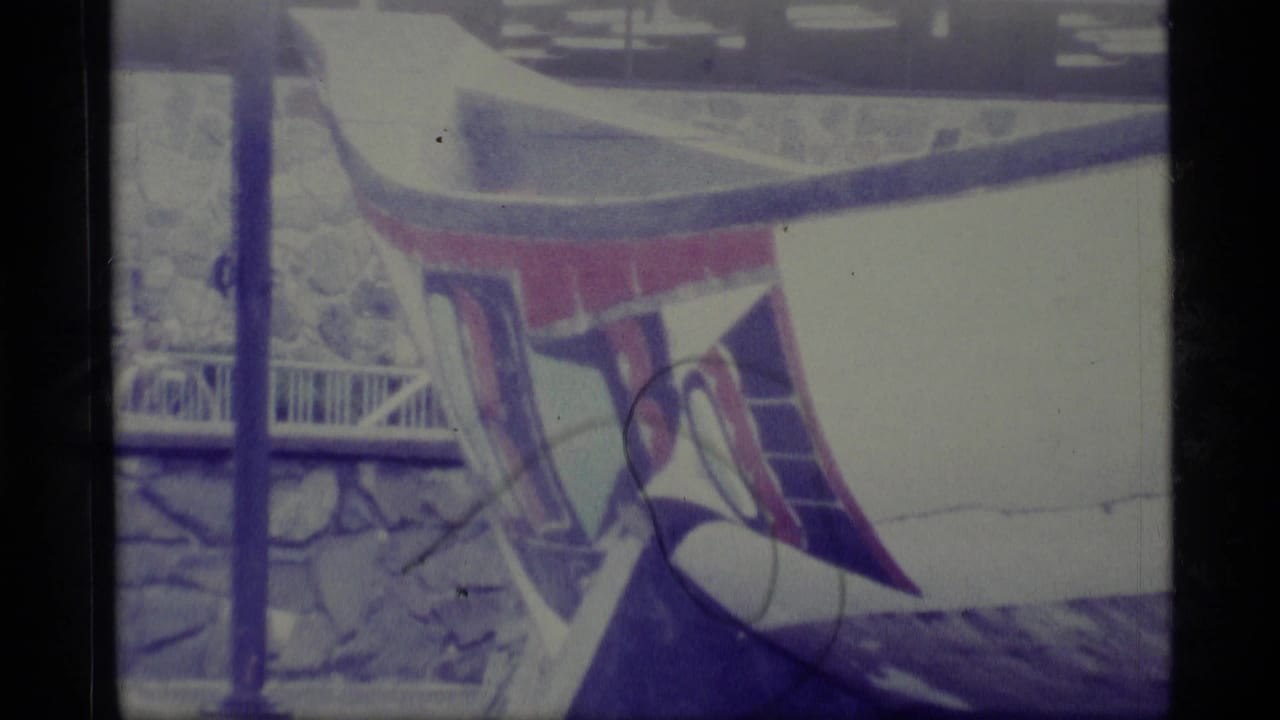 1977 Alaska 8mm frame showing part of a white ceremonial canoe with red and purple painted forms, snow and railing behind it.