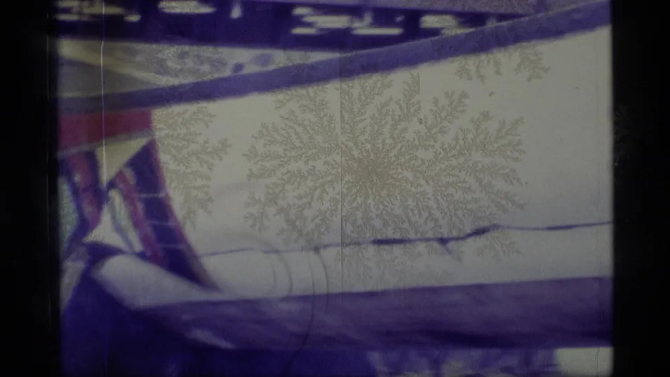 1977 Alaska home-movie frame showing the middle section of a painted canoe on land, with snow, trees and a dark support rail visible.