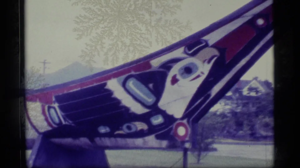 1977 Alaska 8mm frame showing a close, angled view of a painted carved pole with red, black, and white forms against trees and distant mountains.
