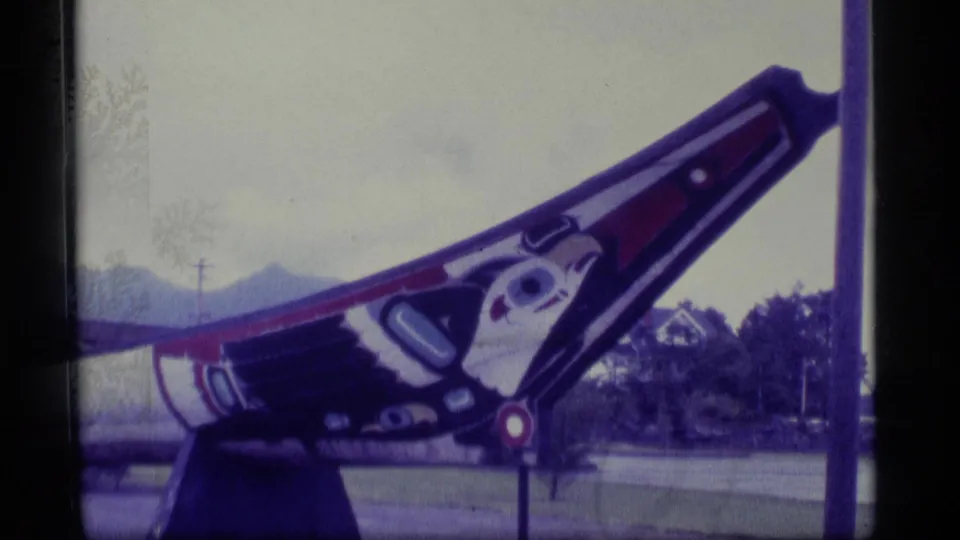 1977 Alaska 8mm frame showing an angled painted carved pole outdoors near a roadway, with trees and low mountains visible beyond.