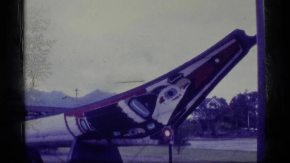 1977 Alaska 8mm frame with a diagonal view of the painted carved pole, cropped close against trees, roadway, and distant mountains.