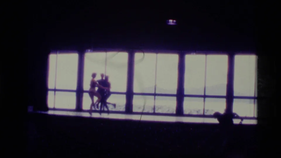 1977 Alaska 8mm frame showing two silhouetted figures on a dim indoor stage, backlit by a wall of large windows.