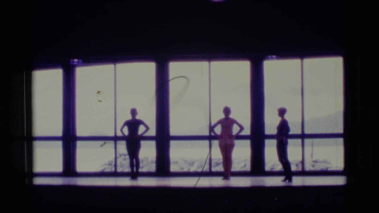 In Alaska 1977 8mm footage, three dancers hold standing poses on a dark stage before large windows overlooking snow and distant hills.