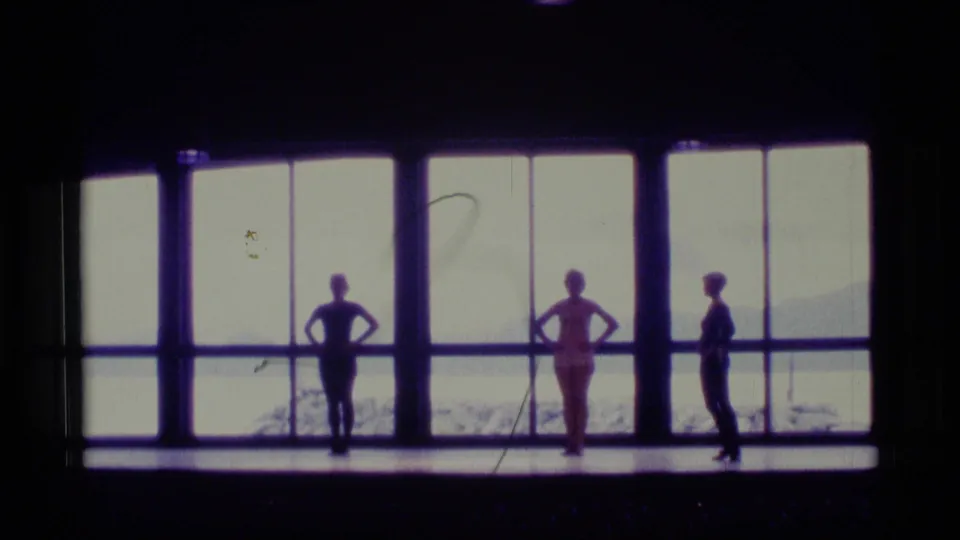In Alaska 1977 8mm footage, three dancers hold standing poses on a dark stage before large windows overlooking snow and distant hills.