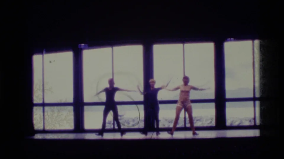 During the 1977 Alaska home-movie clip, three dancers extend their arms in choreography before bright windows and winter scenery.