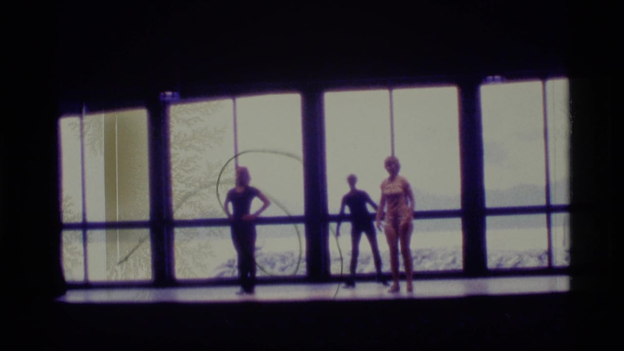 Three dancers in leotards appear in silhouette before large windows in 1977 Alaska home-movie footage, with water and land faintly visible outside.
