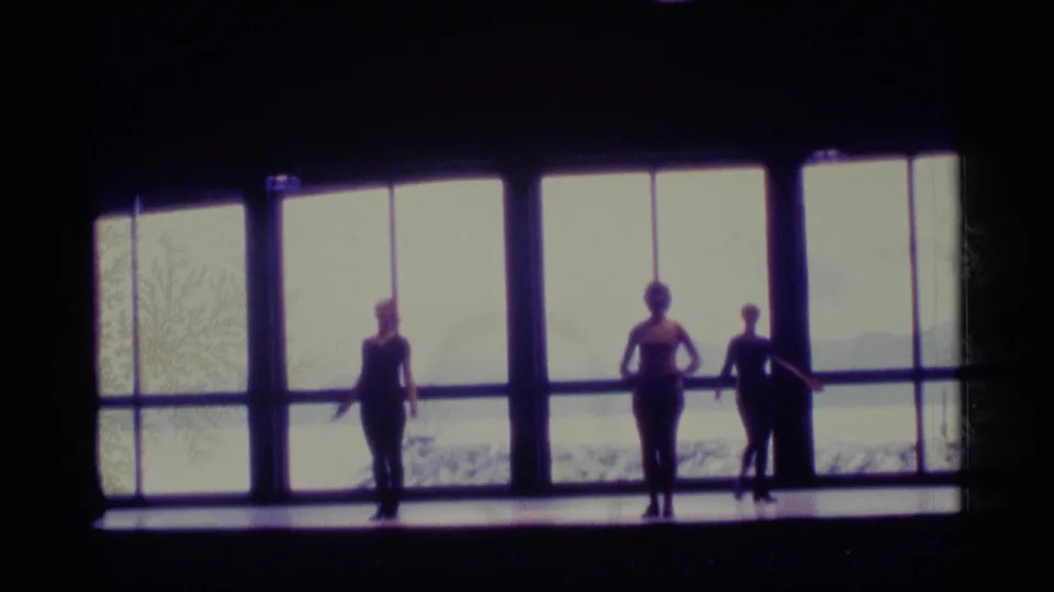 Three dancers are silhouetted against large windows during a 1977 Alaska dance-class scene, with a pale waterfront view behind them.