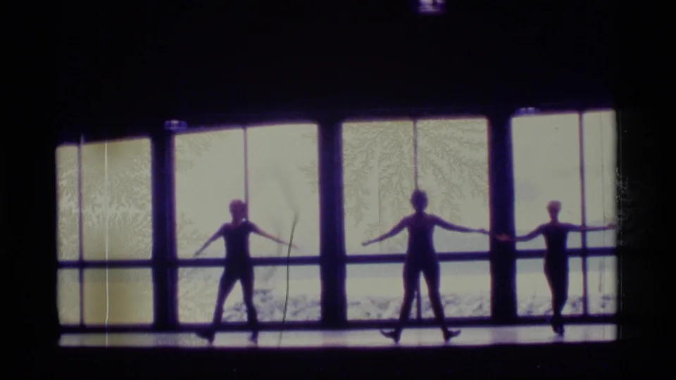 Three dancers hold extended-arm positions in a backlit 1977 Alaska home-movie frame, standing before tall windows and a waterfront view.