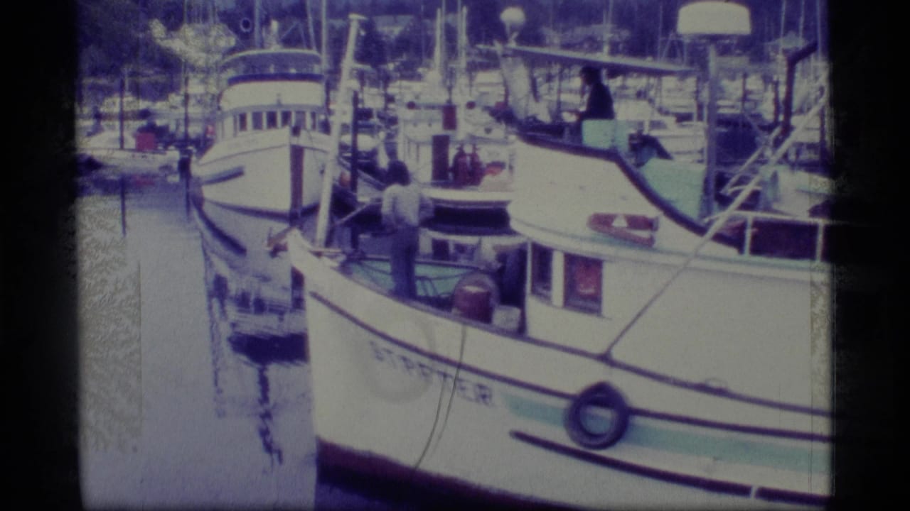 1977 Alaska 8mm frame showing a white fishing vessel in harbor, with men on deck, rigging, dock posts, and boats in the background.