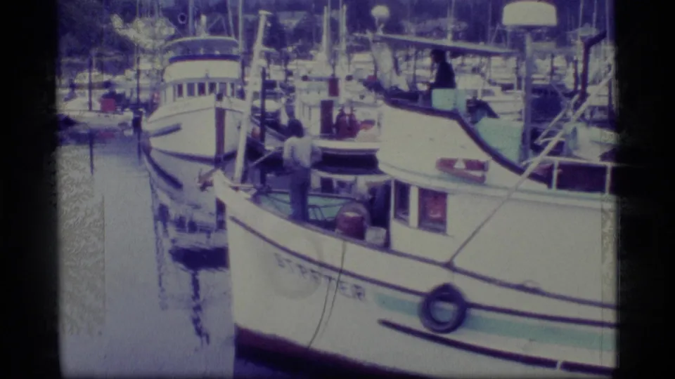 1977 Alaska 8mm frame showing a white fishing vessel in harbor, with men on deck, rigging, dock posts, and boats in the background.