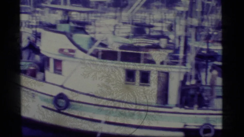 1977 Alaska harbor frame with the side of a white fishing vessel, cabin windows, ropes, masts, and nearby dock structures visible.