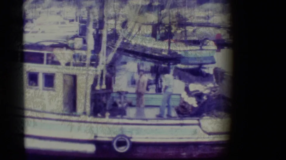 1977 Alaska 8mm frame showing men near deck equipment on a docked fishing vessel, with ropes, a small skiff, masts, and harbor boats around them.