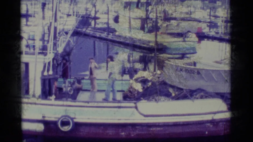 1977 Alaska harbor frame with two men visible on or beside a fishing vessel, plus rigging, ropes, a small boat, and partial hull lettering.
