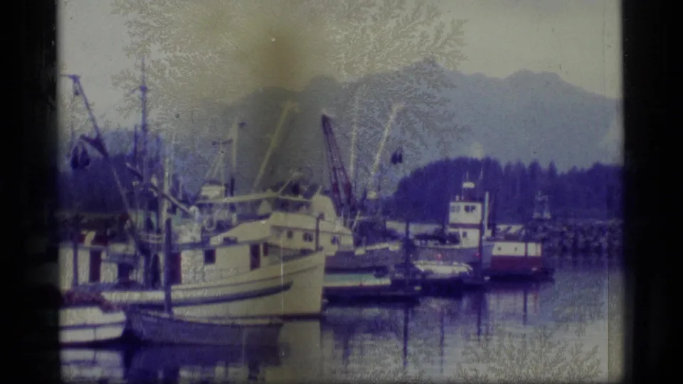 A 1977 Alaska harbor frame shows fishing boats, masts, and calm reflections with a wooded shore and mountain backdrop.
