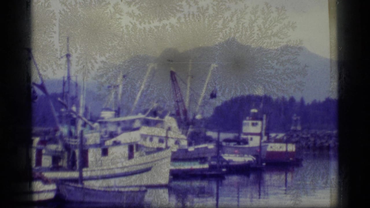 1977 Alaska 8mm frame showing a harbor with moored boats, a larger vessel, calm water, and mountains in the background.