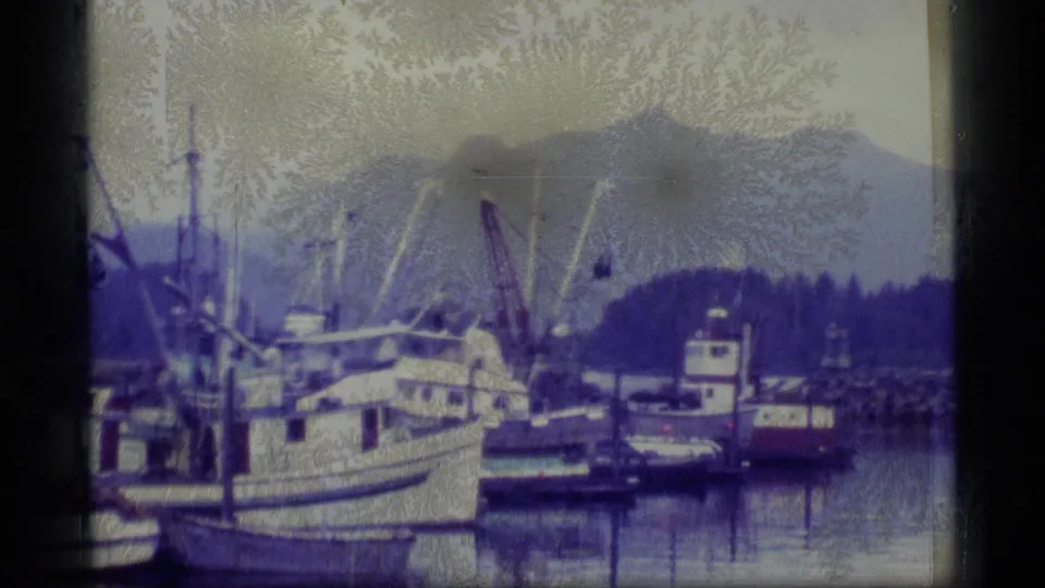 1977 Alaska 8mm frame showing a harbor with moored boats, a larger vessel, calm water, and mountains in the background.