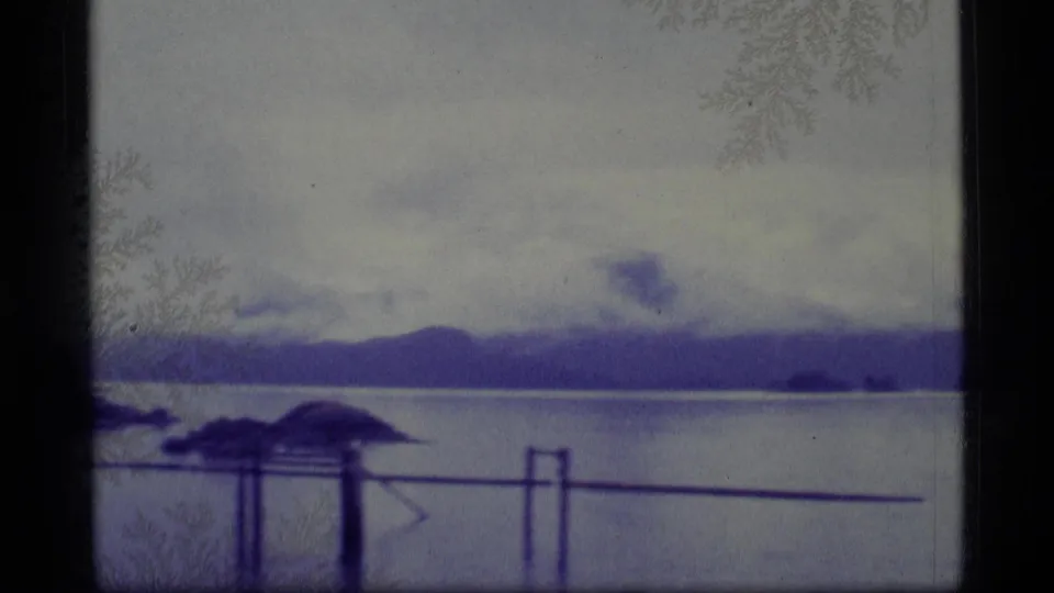 1977 Alaska 8mm frame showing a quiet waterway, low island, distant hills, cloudy sky, and dark dock railings near the camera.