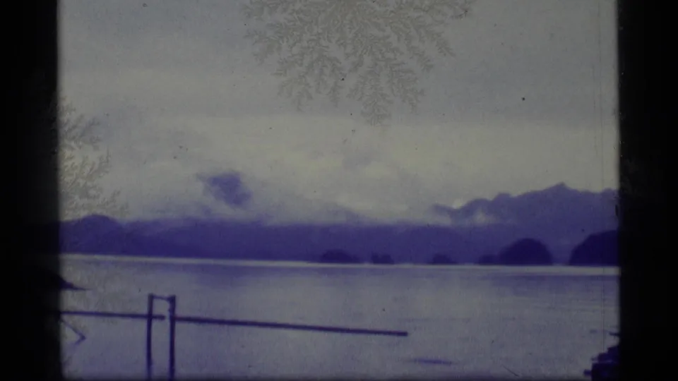 1977 Alaska 8mm frame with open calm water, distant hills, cloudy sky, and a simple railing crossing the lower foreground.