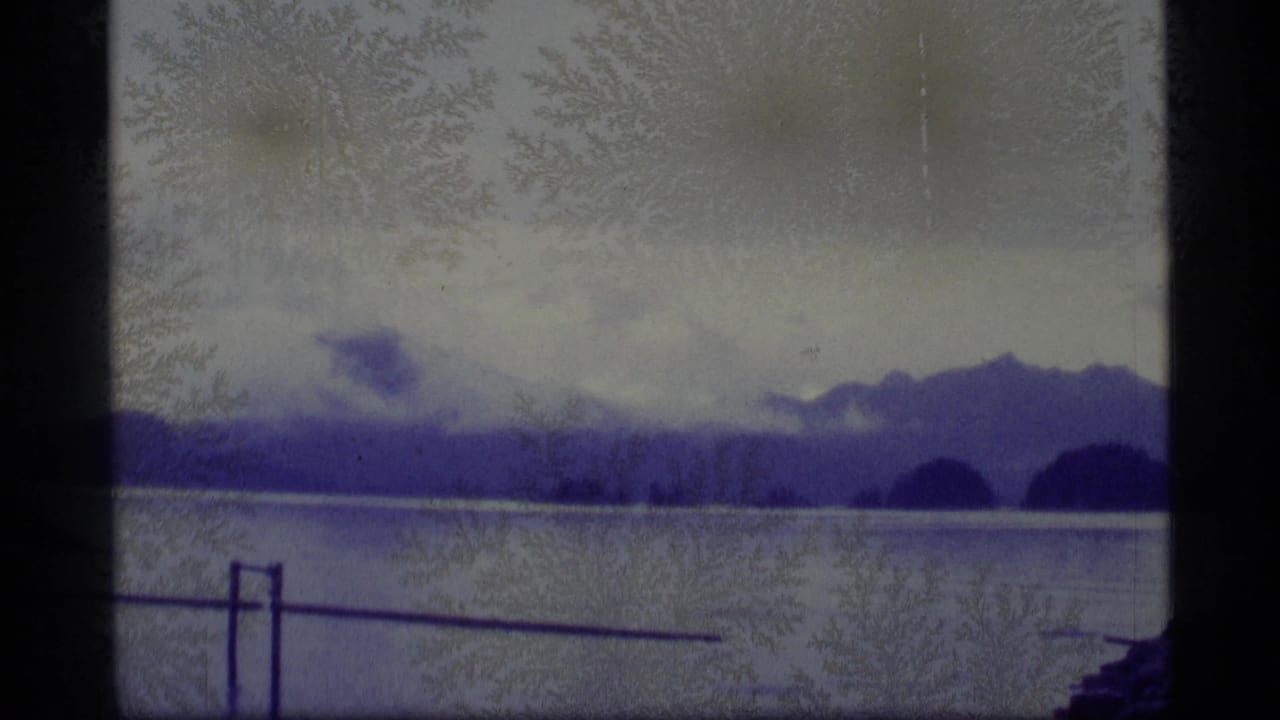 1977 Alaska 8mm frame showing calm lake water, distant mountain silhouettes, cloudy sky, and a foreground railing with aged-film artifacts.